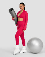 Elevate Compression Long Sleeve Jacket in Strawberry, front view zipped, holding a yoga mat, exercise ball beside her